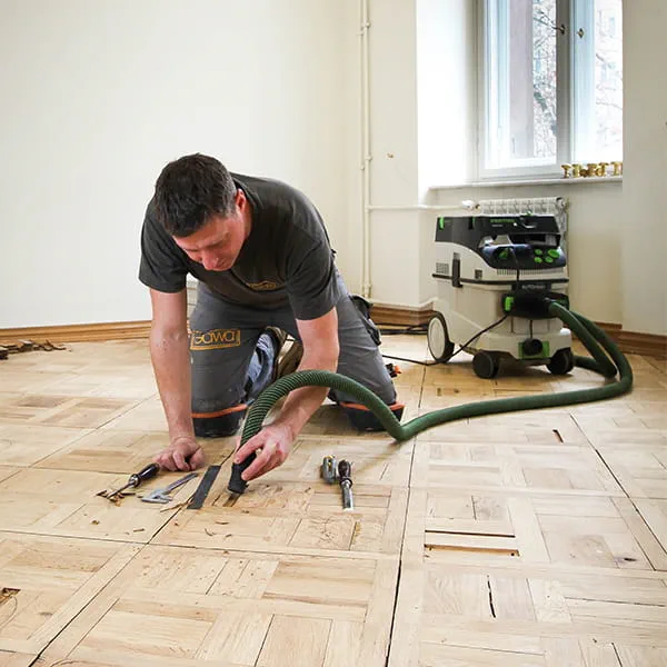 Mistrz parkieciarstwa Jarek Gawina odrywa uszkodzone elementy od parkietu i usuwa je za pomocą mobilnego ssaka Festool